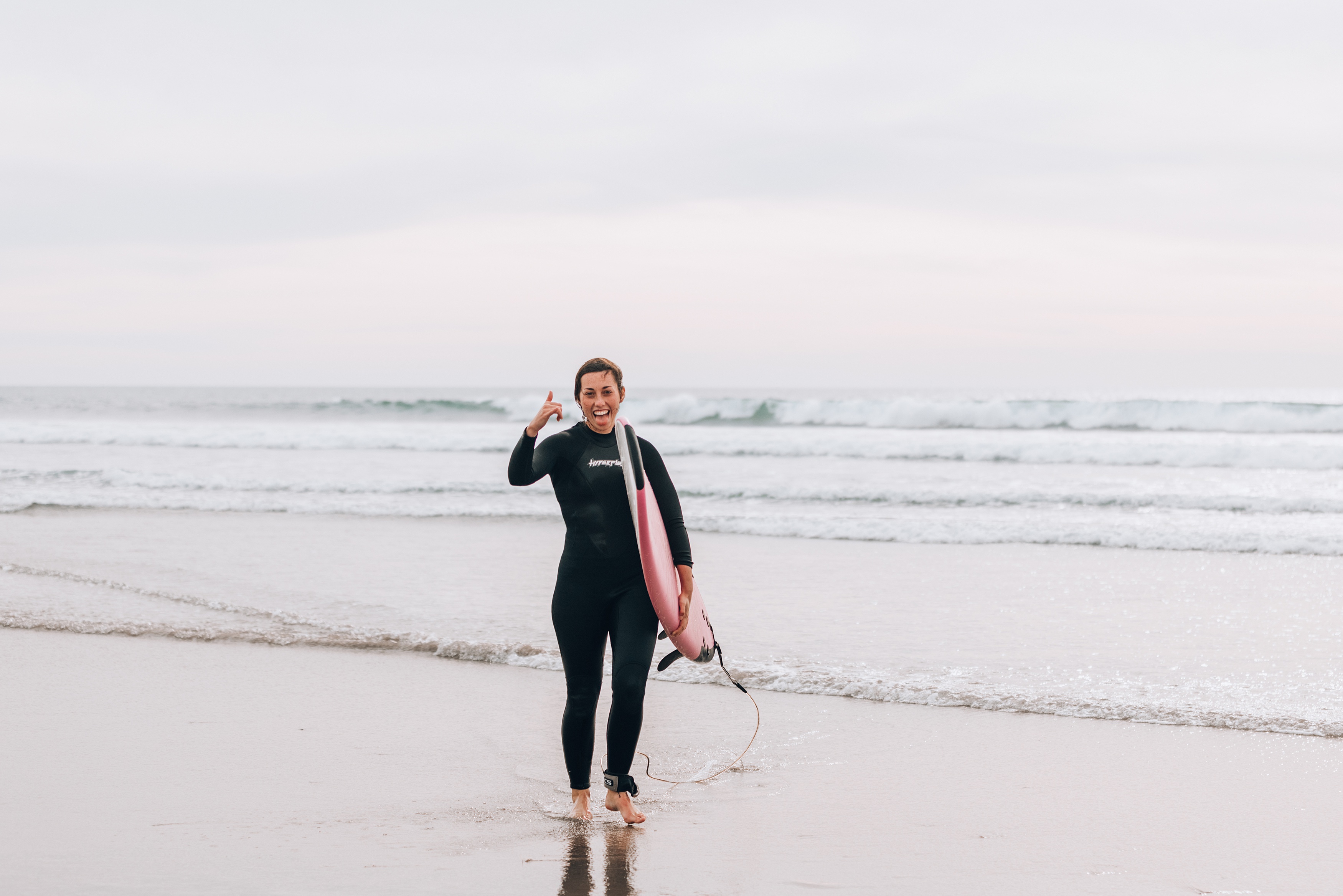 girl walking on beach in wetsuit with surfboard